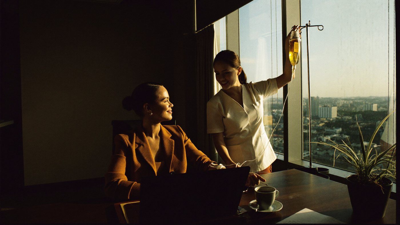 A registered nurse setting up a vitamin IV drip for a professional at her high-rise office desk